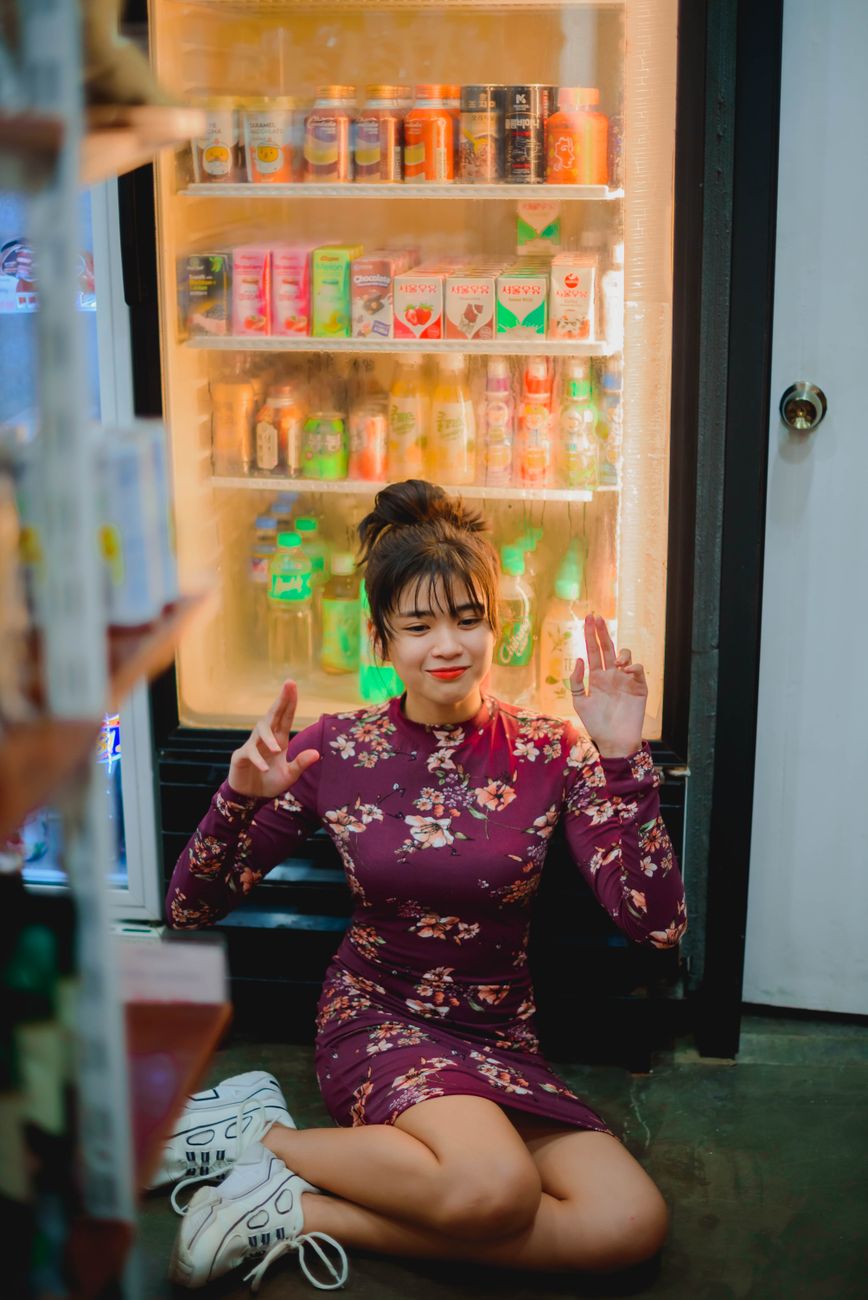 Asian woman in a floral dress sits playfully in front of a beverage fridge indoors.