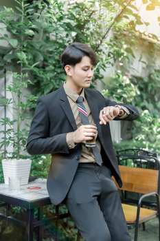 Young man in formal suit outdoors checking wristwatch while holding a glass.