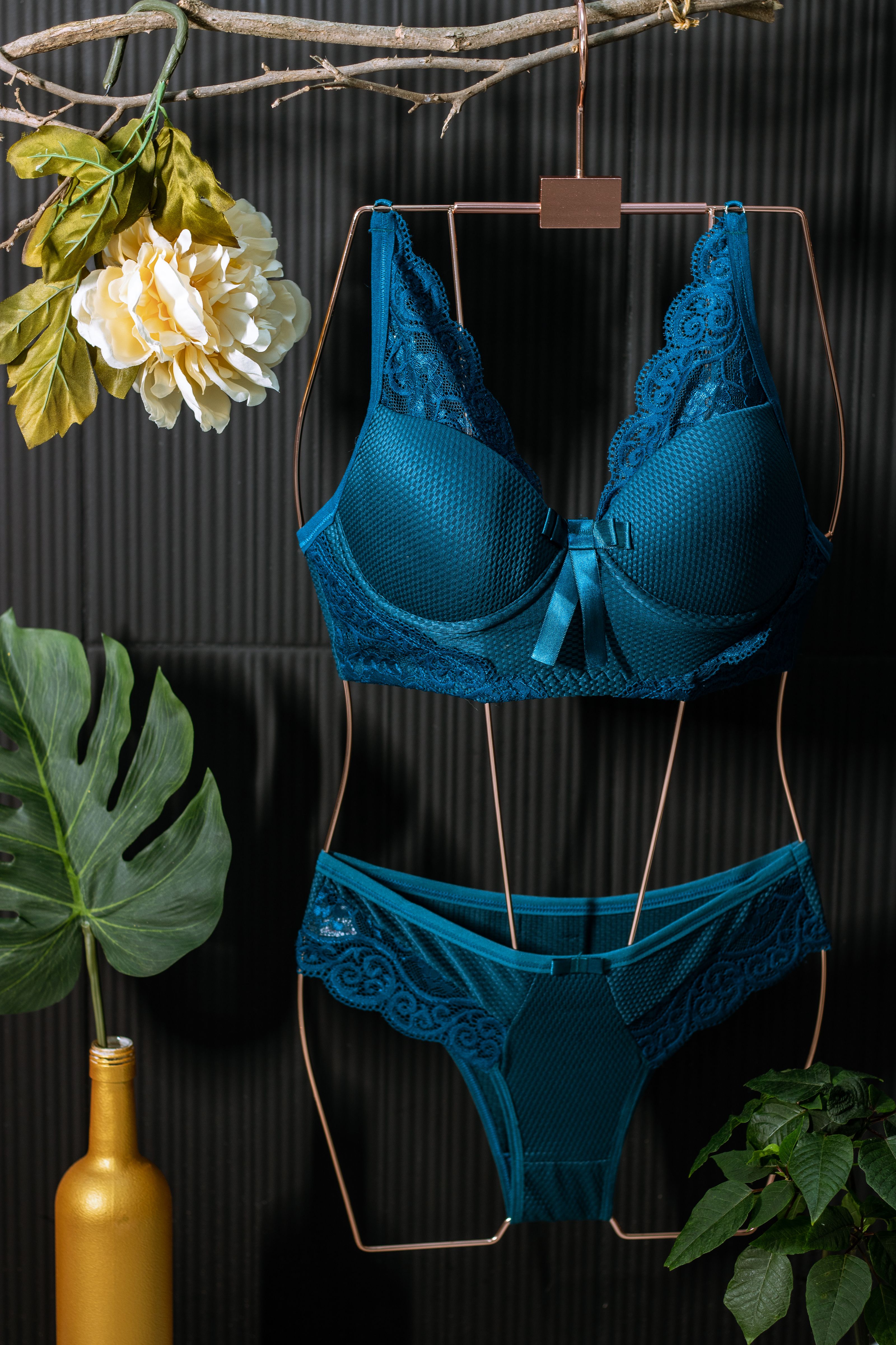 Conjunto de lingerie azul em cabide com folhas e flores decorativas contra fundo escuro.