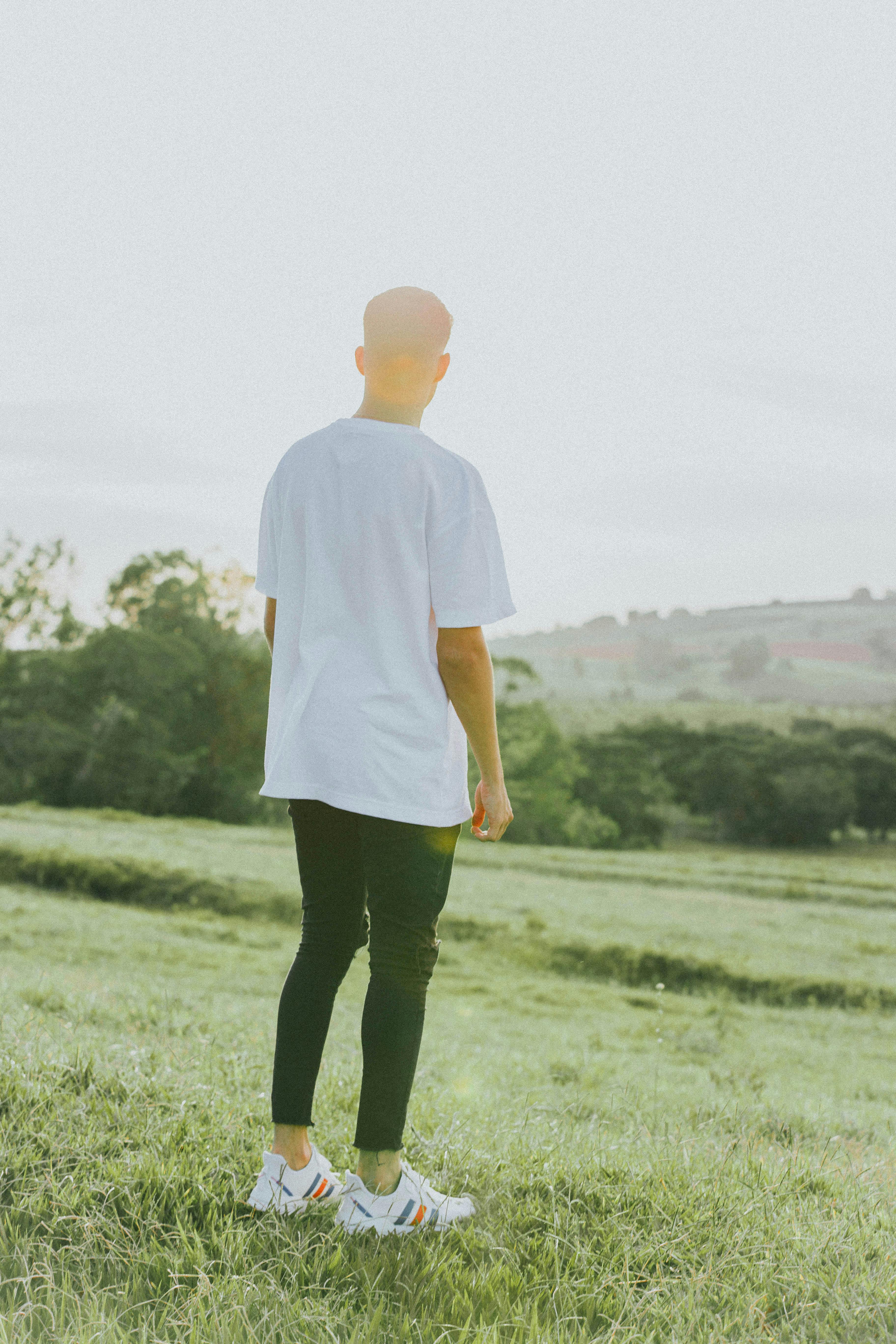 A man stands in a green field wearing a white shirt and black pants, enjoying the serene landscape.