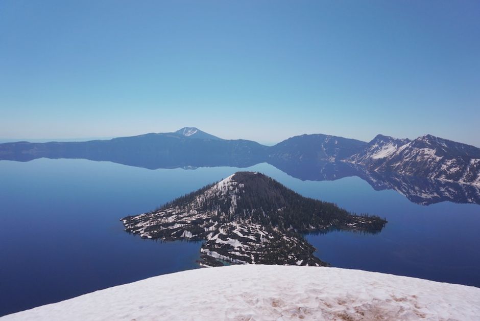 Scenic view of Crater Lake's calm waters reflecting snow-capped peaks under a clear blue sky.