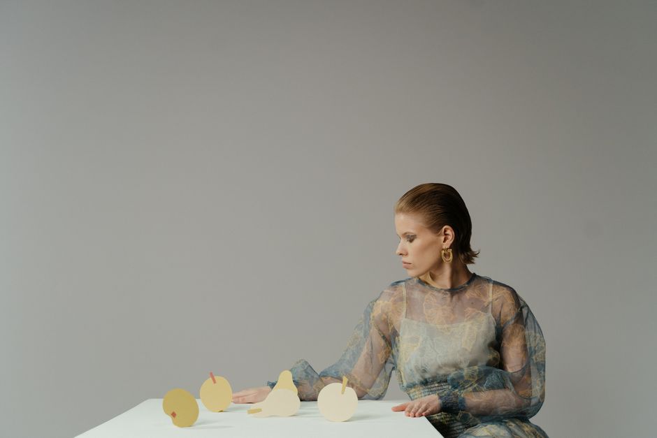 Conceptual image featuring a woman in a sheer dress with papercraft fruits on a table.