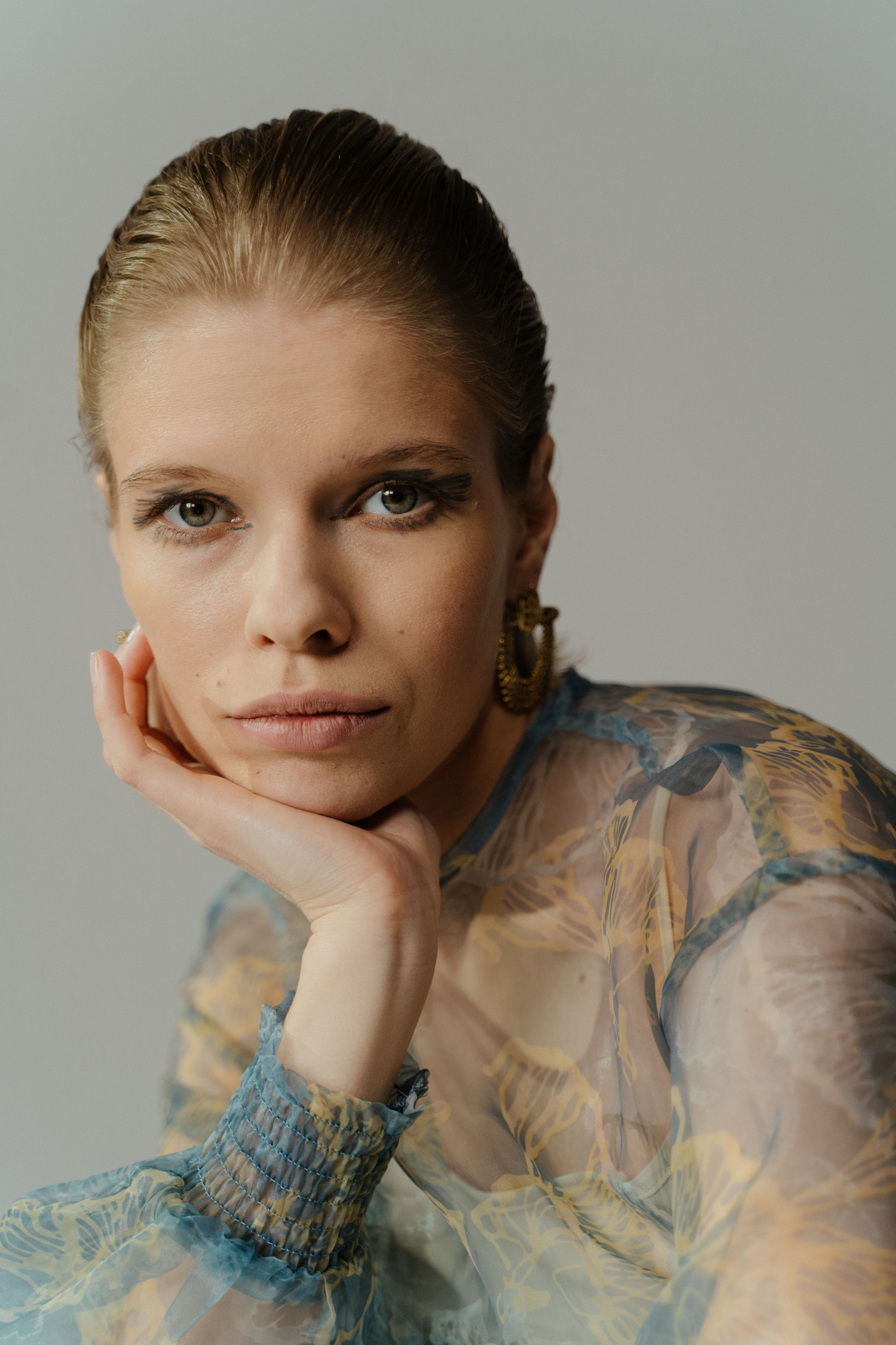 Close-up portrait of a woman with hand on chin, wearing a sheer blouse and earrings.