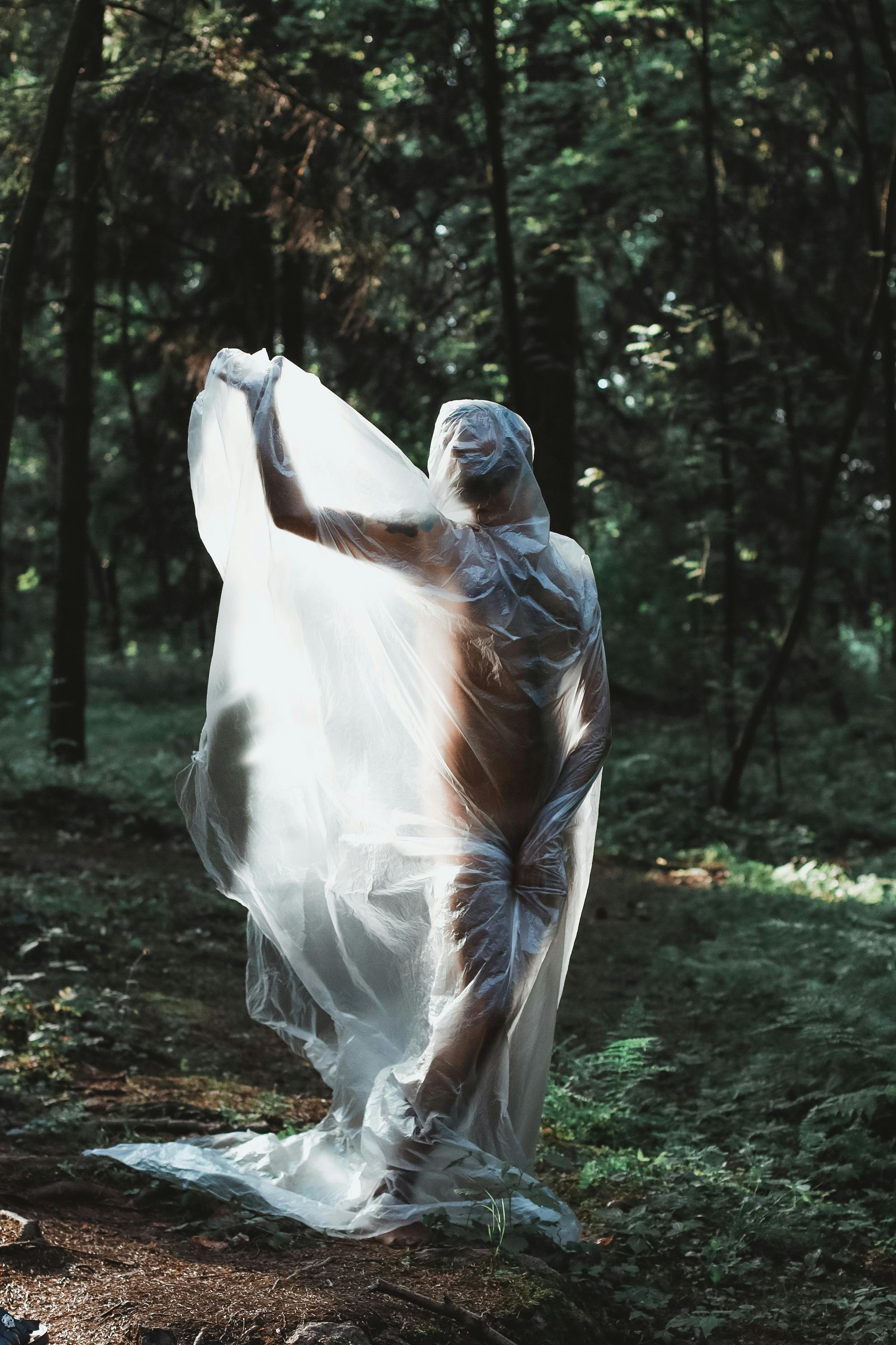 A man wrapped in sheer plastic stands in a sunlit forest, creating a contrast between nature and artificiality.