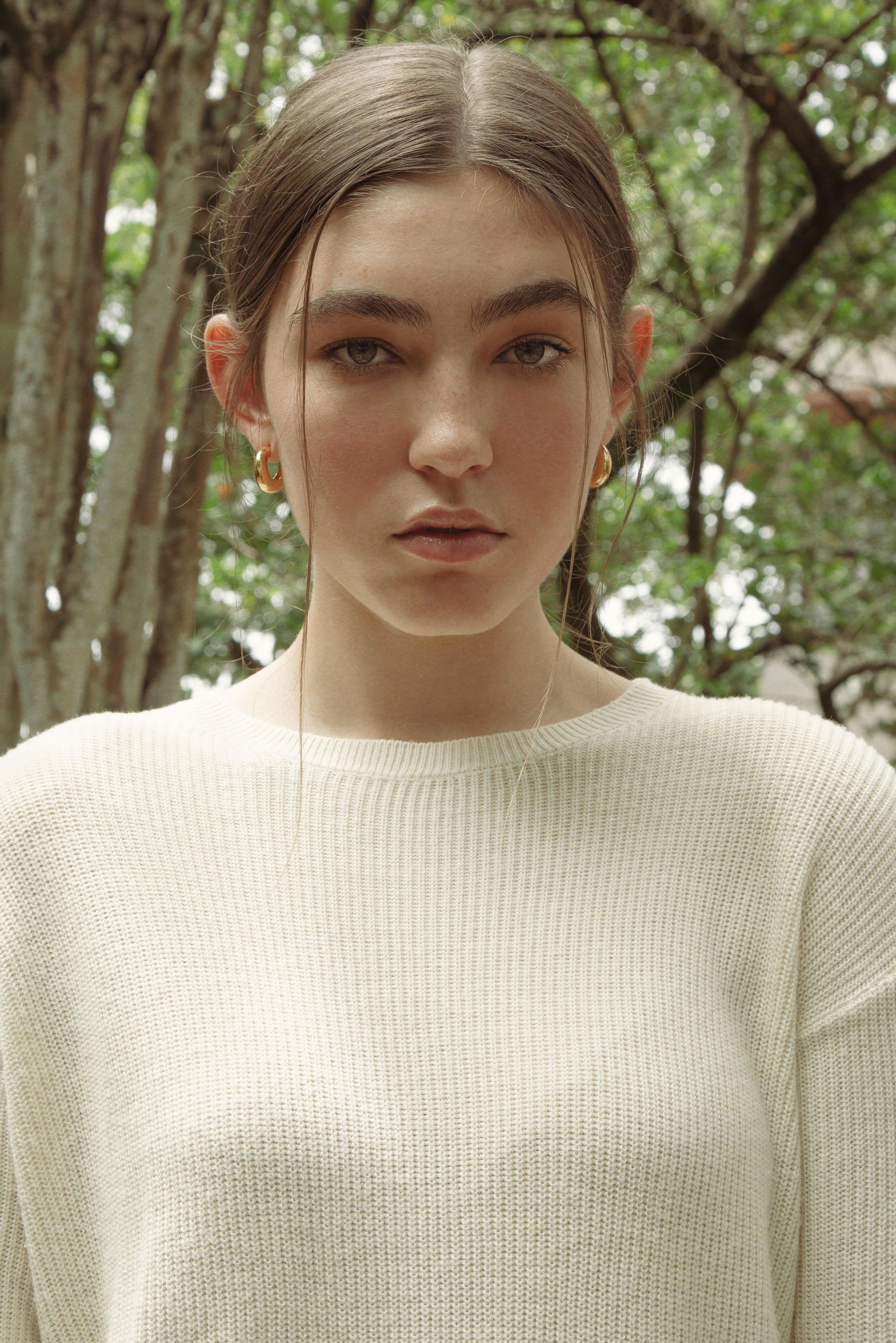 Close-up portrait of a young woman in a knitted sweater with loop earrings, standing outdoors.