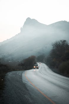 A scenic road through misty mountains at dusk with cars driving amidst foggy conditions.