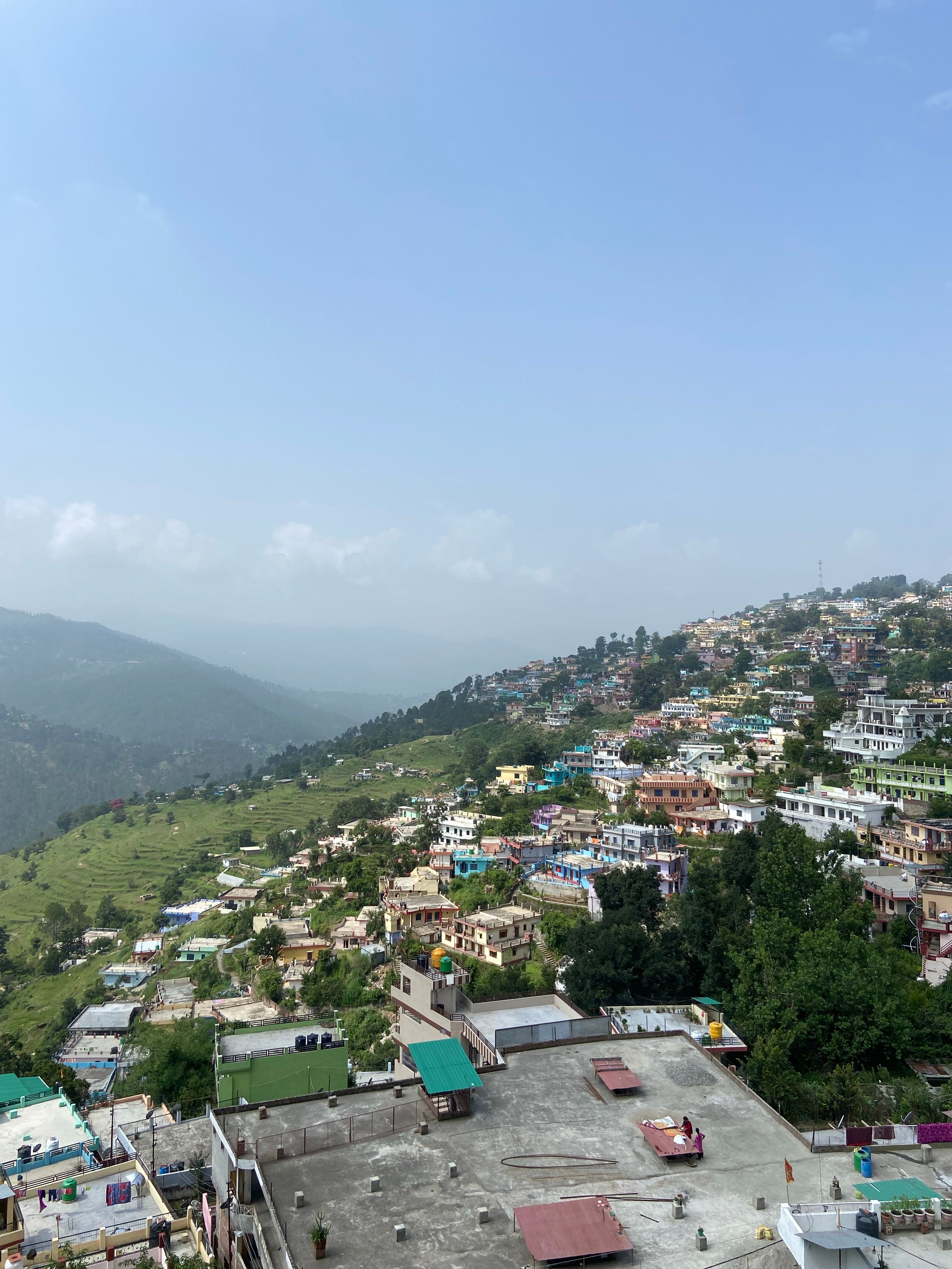 Aerial view of a vibrant hillside town in India with lush greenery and scattered buildings under a blue sky.