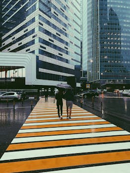 Pedestrians with umbrellas cross a colorful crosswalk on a rainy day in Moscow's modern cityscape.