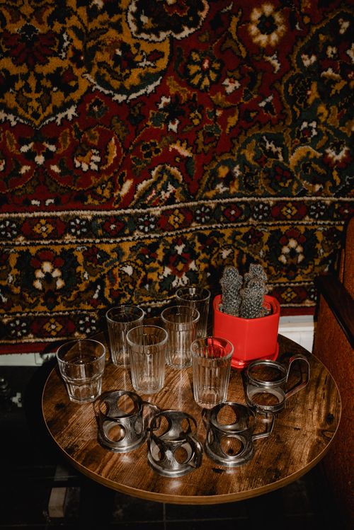 Free A cozy still life setup featuring a wooden table with vintage glasses and a cactus against a floral tapestry backdrop. Stock Photo