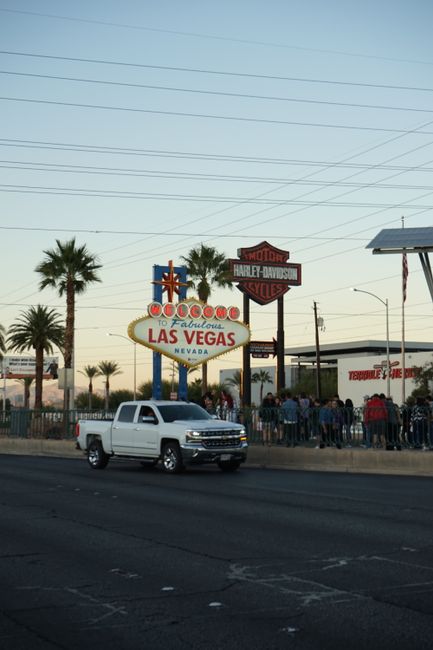 Classic Las Vegas sign with a Chevrolet Silverado passing by and a crowd. Palm trees line the road.