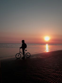 A person stands with a bicycle on a beach at sunrise, creating a serene silhouette against the sea.