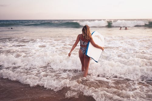 Woman in Blue and White Bikini Holding White and Blue Surfboard Walking on Beach