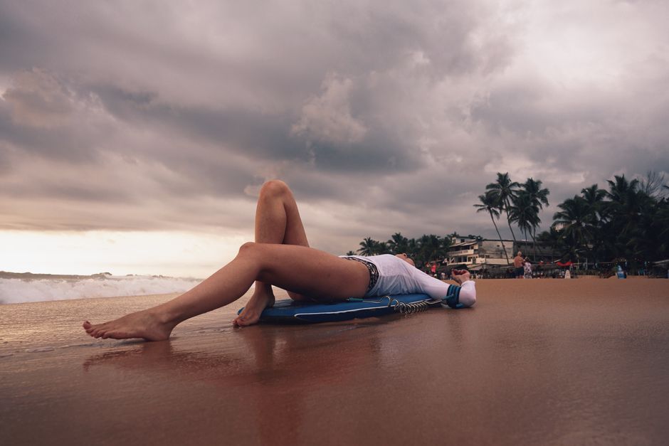 Woman in swimwear relaxing with a surfboard on a sandy beach beneath a cloudy sky.