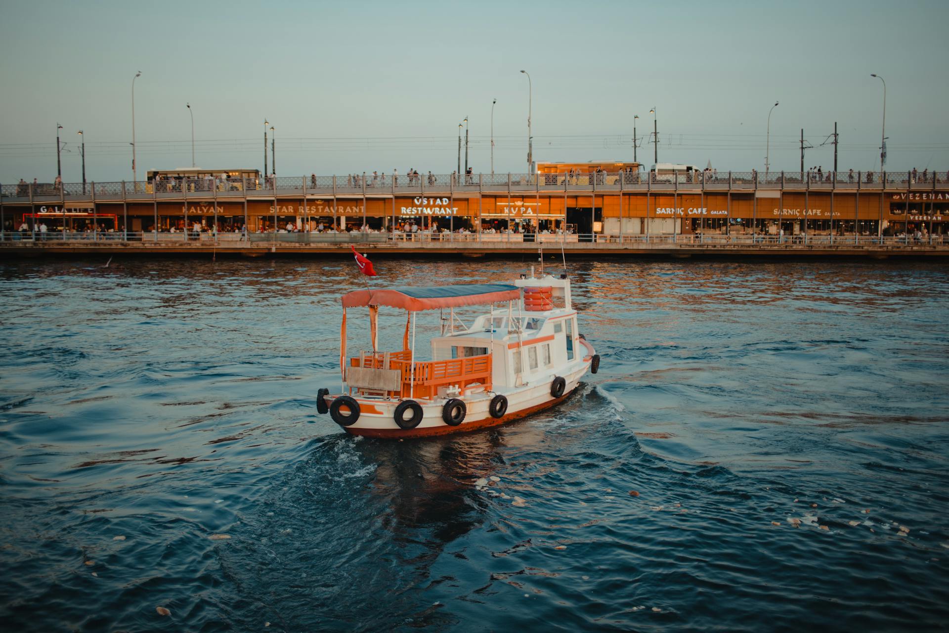A scenic view of a tour boat sailing on the Golden Horn in Istanbul with a backdrop of bustling waterfront restaurants.