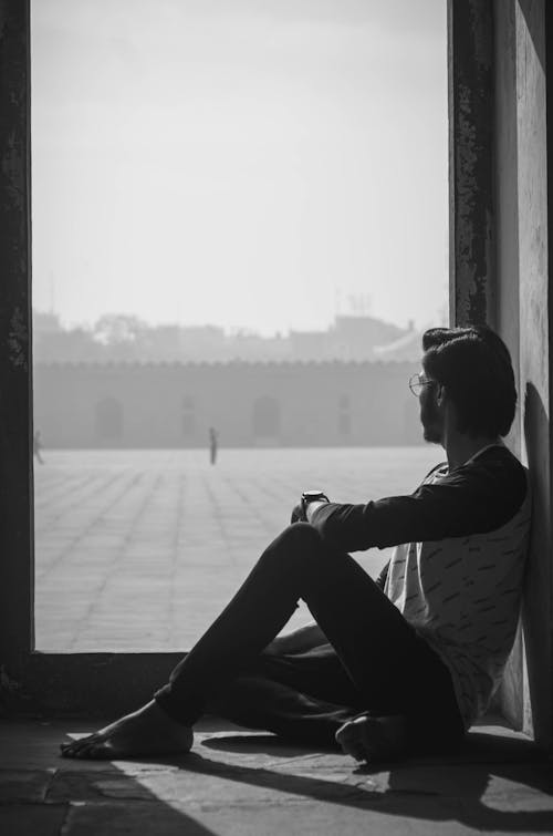 Grayscale Photo of Man Sitting on the Floor While Looking Outside 