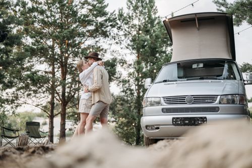 Free A couple shares an intimate moment near a camper van in a picturesque forest campsite. Stock Photo