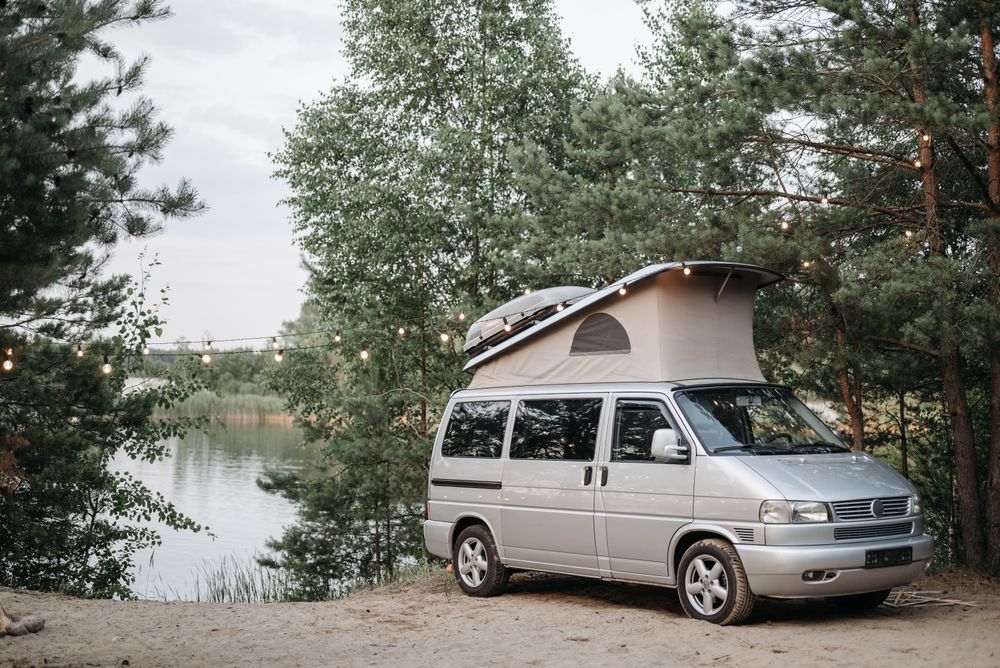 Campervan mic cu acoperiș ridicabil, parcat lângă un lac liniștit