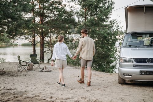 Free A couple enjoys a scenic lakeside camp with a camper van and trees under string lights. Stock Photo