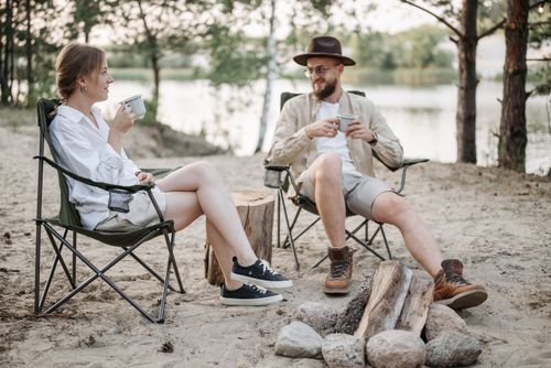 Free A couple enjoys a peaceful camping experience by the lake, sitting in camping chairs with drinks in hand. Stock Photo