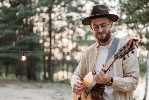 Free Bearded man with a fedora playing acoustic guitar in a serene outdoor setting. Stock Photo
