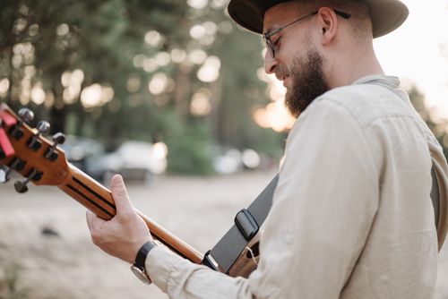 Free A bearded man playing an acoustic guitar outdoors while wearing a fedora hat, captured in a warm setting. Stock Photo