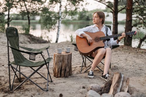 Free A woman sitting by a lake playing guitar, surrounded by nature and camping gear. Stock Photo