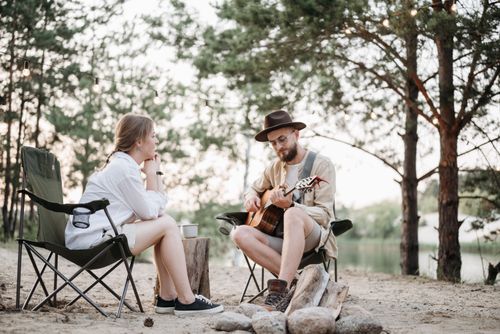 Free A couple enjoying a peaceful day outdoors, with a man playing guitar by a lakeside campsite. Stock Photo