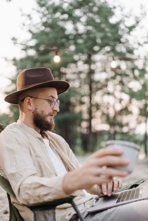 Free A man with a beard and hat uses a laptop outdoors, enjoying a coffee. Ideal for digital nomad themes. Stock Photo