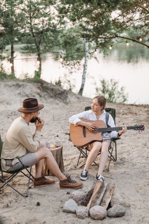 Free A couple enjoying a musical evening by the campfire, playing guitar and harmonica at a lakeside campsite. Stock Photo