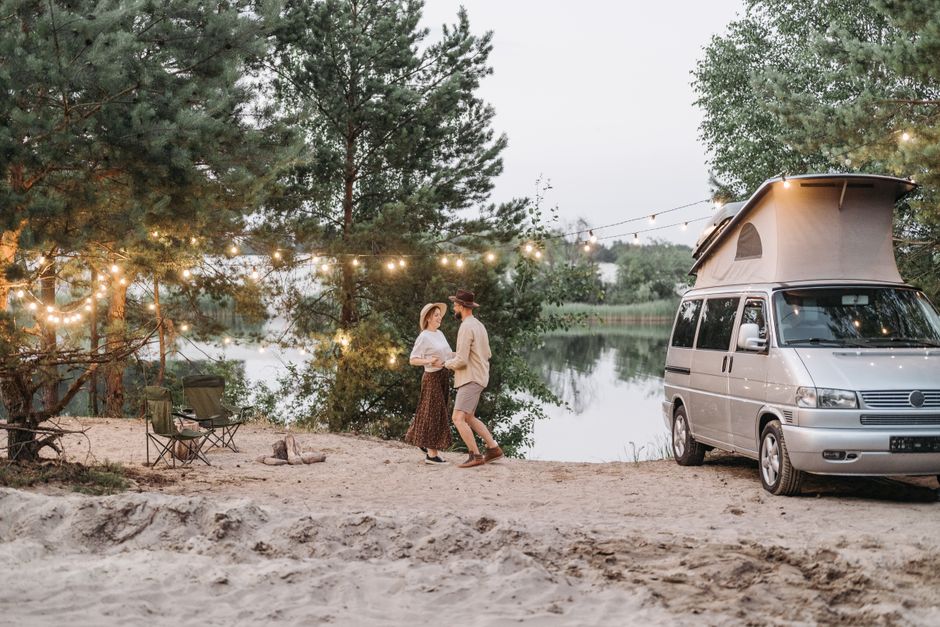 Couple dancing by a camper van under string lights near a serene lake, enjoying a peaceful camping evening.