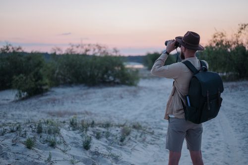 Free Man with backpack observing nature through binoculars during a scenic sunset. Stock Photo