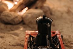 A cozy campfire scene with a moka pot brewing coffee on a portable stove in sandy outdoors.