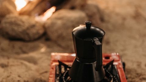 A cozy campfire scene with a moka pot brewing coffee on a portable stove in sandy outdoors.