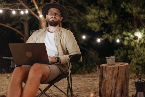Man Sitting on a Folding Chair on a Beach at Night and Using a Laptop 