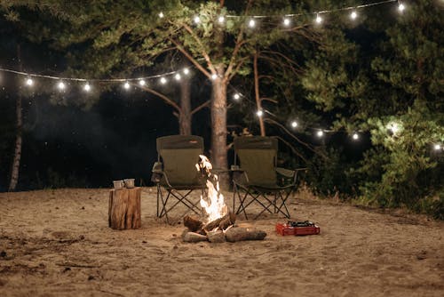 Folding Chairs Standing next to a Bonfire on a Beach with Sting Lights hanging between the Trees