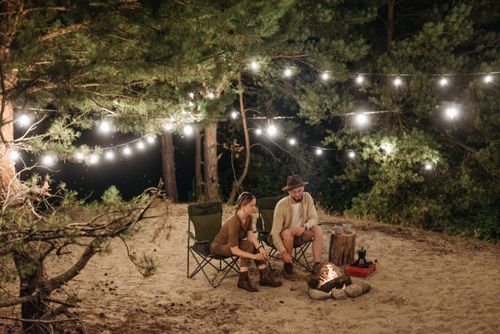 Free Couple enjoying a quiet night camping with a campfire and string lights in a forest setting. Stock Photo
