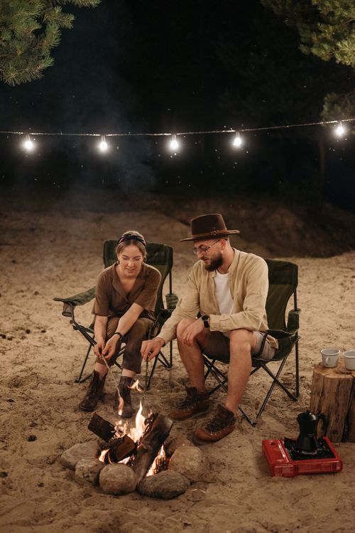Free A couple enjoys a cozy evening by a campfire on a sandy beach, surrounded by string lights. Stock Photo