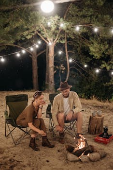 A couple relaxes by a campfire under string lights, enjoying a serene camping night.