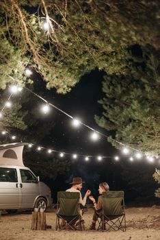 A couple enjoys a quiet night camping under string lights by a van amidst green trees.