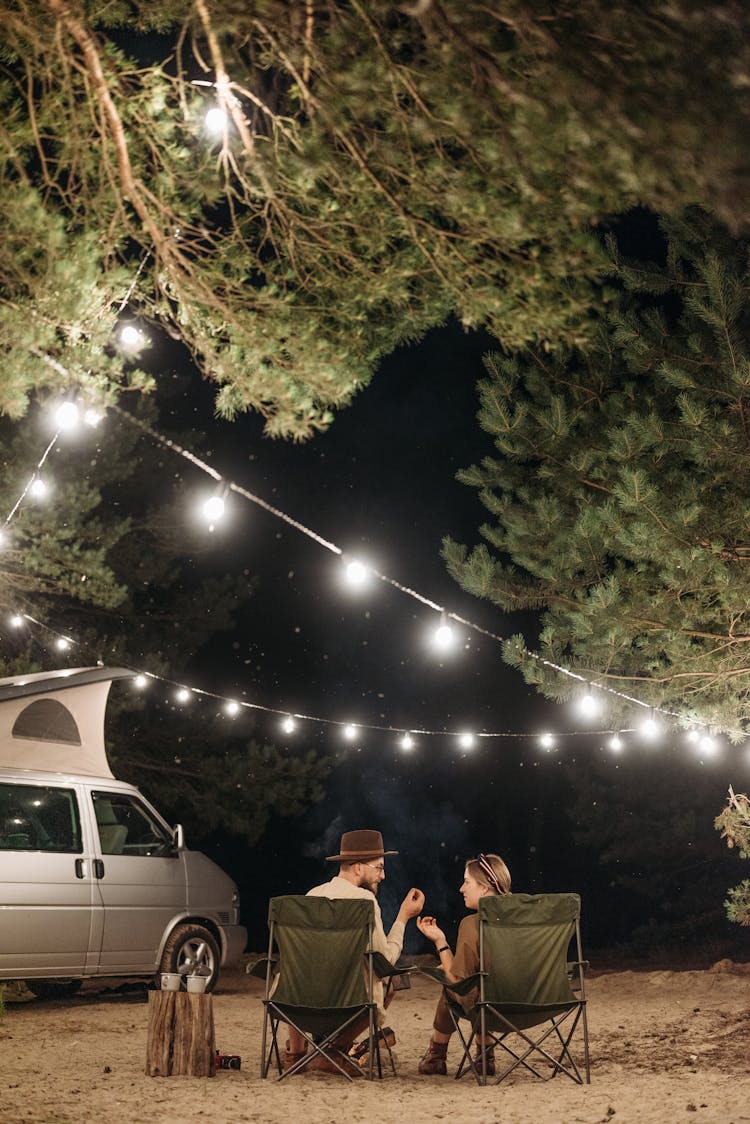 A Couple Sitting On The Folding Chair While Having Conversation Under The Green Trees At Night