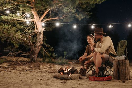 Free Couple enjoying a cozy camping night by a bonfire under festive lights. Stock Photo