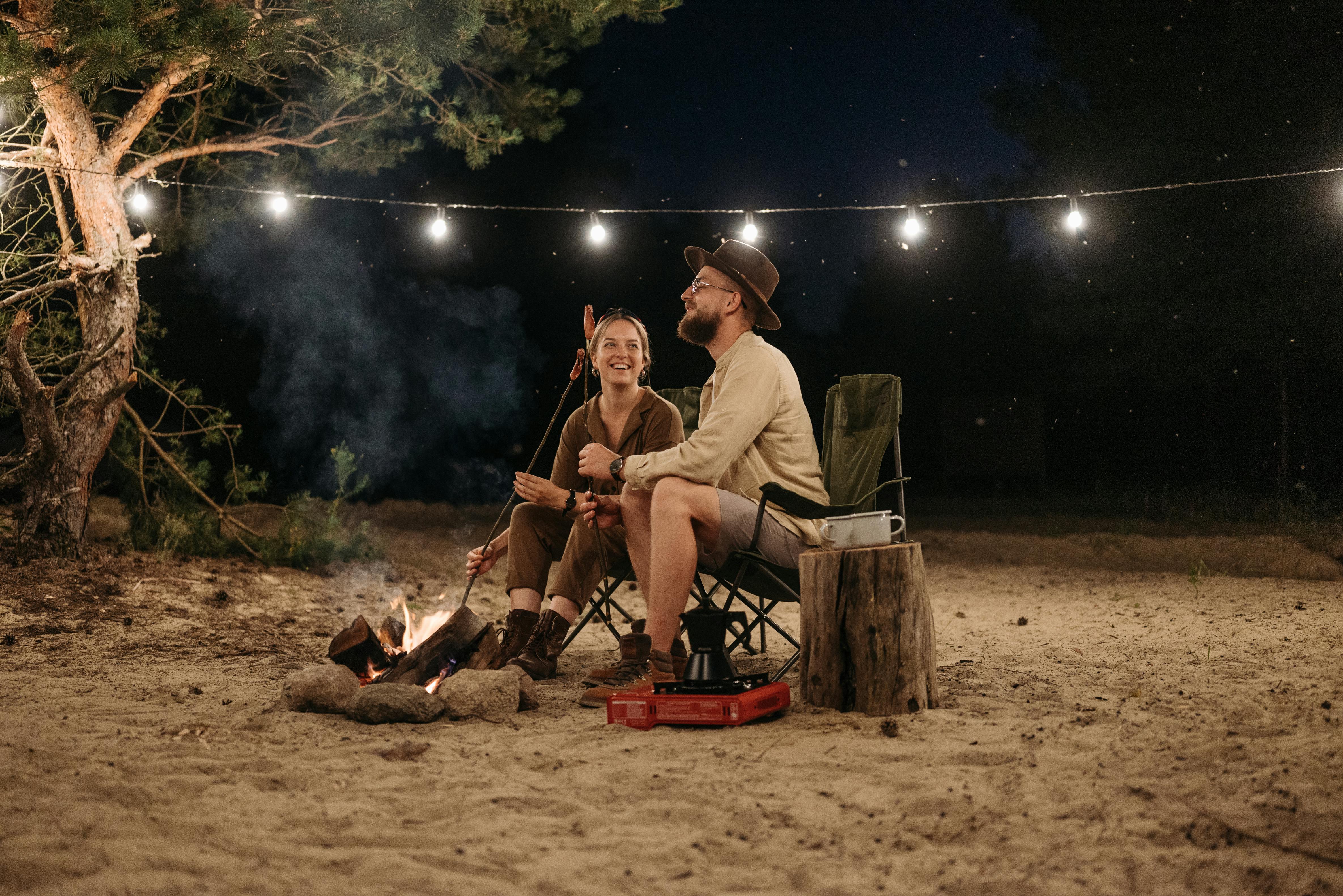 a couple sitting on chair while grilling on campfire