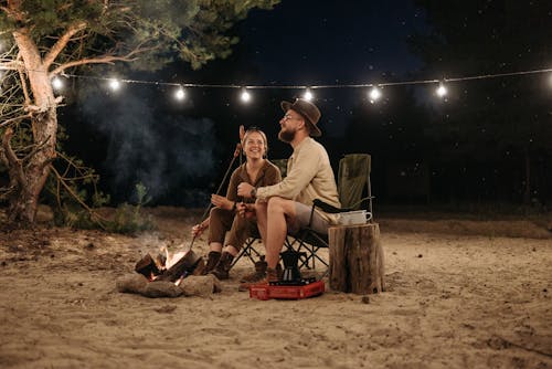 A Couple Sitting on Chair while Grilling on Campfire