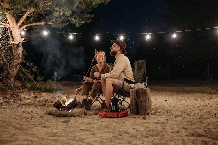 A Couple Sitting On Chair While Grilling On Campfire