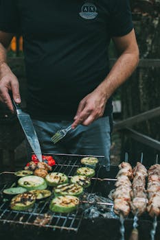 A man grilling vegetables and meat skewers on an outdoor barbecue.
