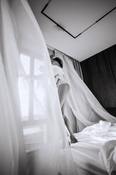 Black and white photo of a woman in lingerie standing by a window with flowing curtains.