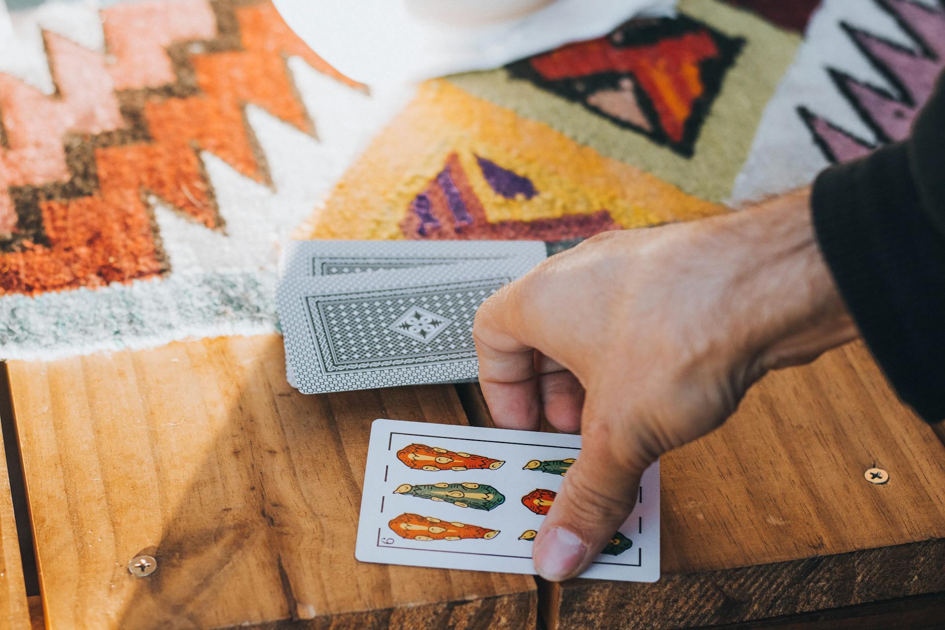 Close-up of a hand dealing Spanish playing cards on a wooden table with a colorful textile background.