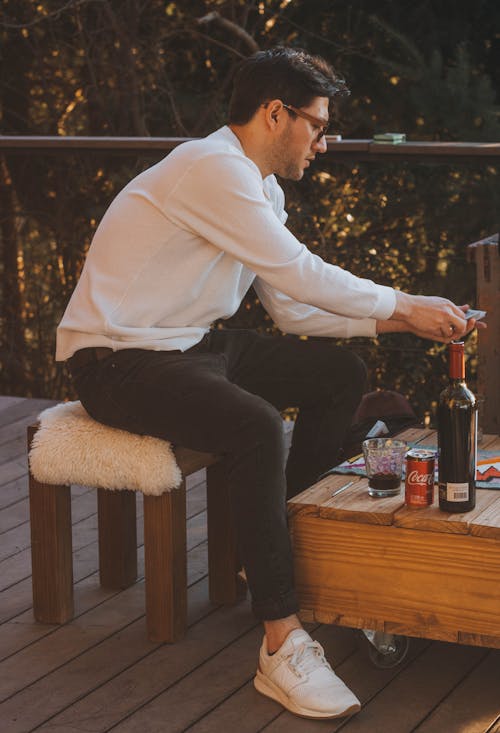 A Man in White Long Sleeves and Black Pants Sitting Near the Wooden Table with Wine Bottle