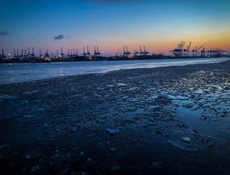 日落时分，工业港口一片繁忙，起重机的剪影映在冰冷的水面上