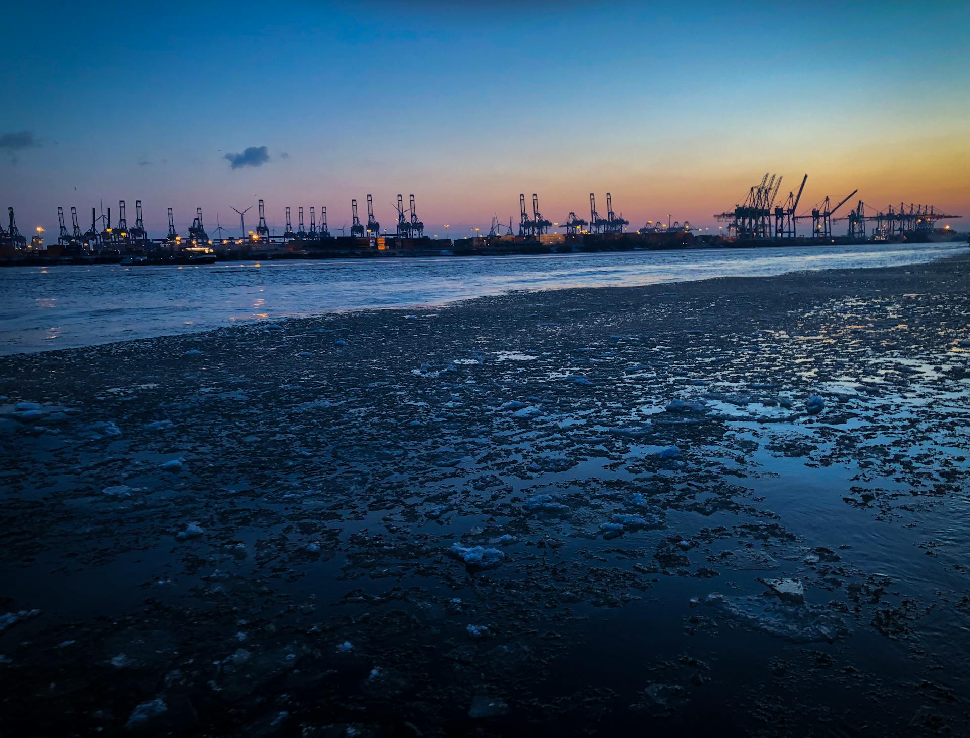 Silhouetted cranes at a bustling industrial port during sunset, with icy water in the foreground.